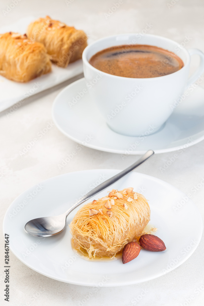Greek pastry Kataifi with shredded filo dough stuffed with almond nuts, in honey syrup, on a white plate, served with cup of coffee, vertical