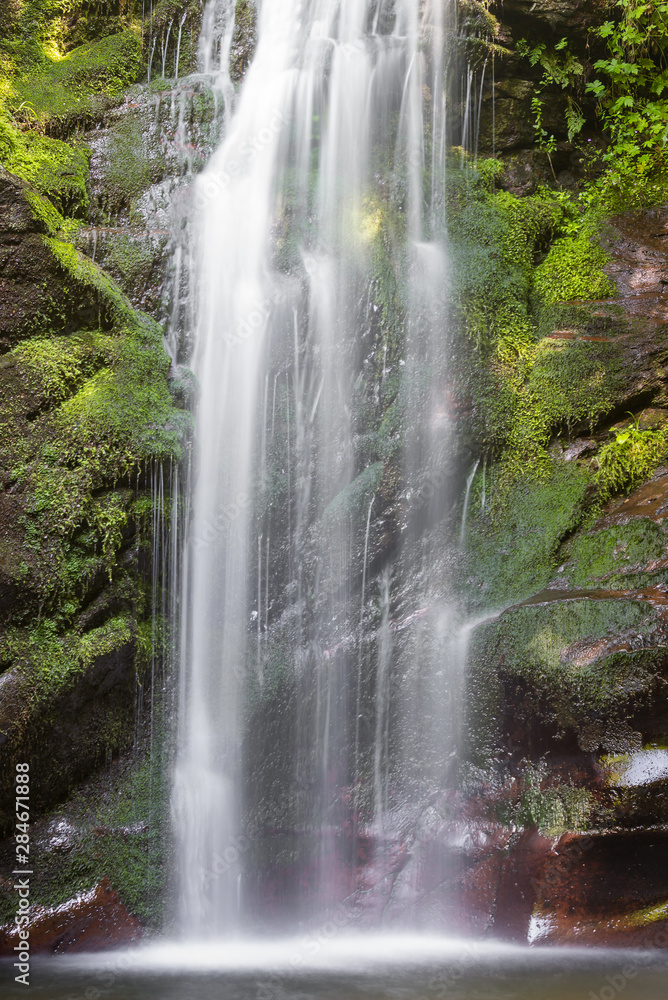 Obraz premium Scenic mountain waterfall cascading down the rocky cliff covered by colorful, vivid green moss and patches of sun hitting the rocks