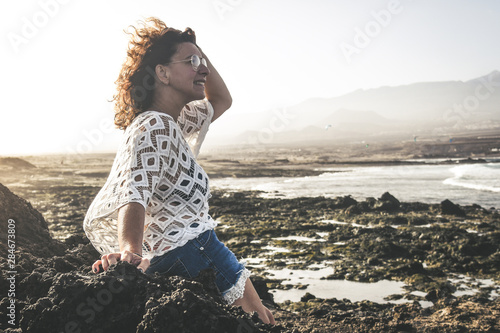 Trendy woman sitting on the rocks enjoy summer holiday outdoor. Portrait of smiling girl with glasses. Wind blown in the hair. Happy fashion female tourist. Tourism freedom and carefree concept. photo