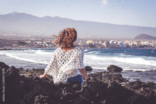 Back view of a Trendy woman sitting on the rocks enjoy summer holiday outdoor. Portrait of a girl relaxing. Wind blown in the hair. Happy fashion female tourist. Tourism freedom and carefree concept. photo