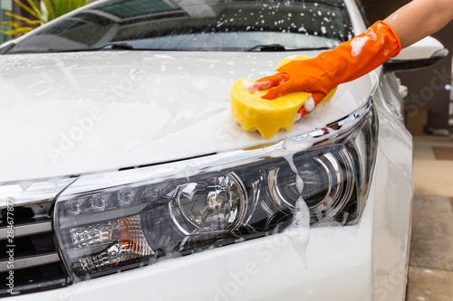 Woman hand wearing orange gloves with yellow sponge washing headlight modern car or cleaning automobile. Car wash concept