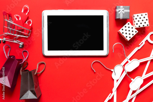 Creative top view flat lay online shopping tablet, trolley with packages, gift boxes. Ecommerce concept, christmas shopping on red background