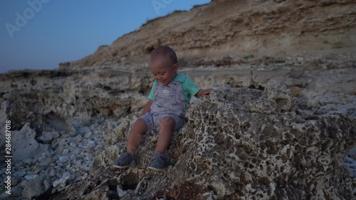 Kid sitting on the stones on the sea coast at sunset . Boy talking to himself and fidgeting