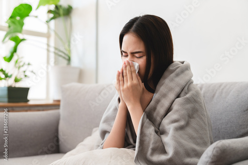 Valokuvatapetti sick asian young woman sneezing into tissue paper while covered with a blanket a