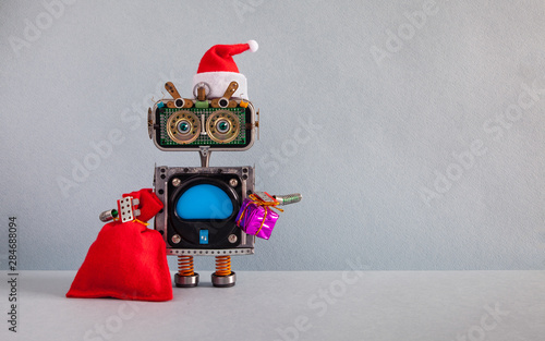 Xmas New Year Santa Claus robotic greeting card. Happy robot in Santa hat, red bag gift and purple box on gray wall background. Copy space for New Year data or Ho Ho Ho on blue screen body. photo