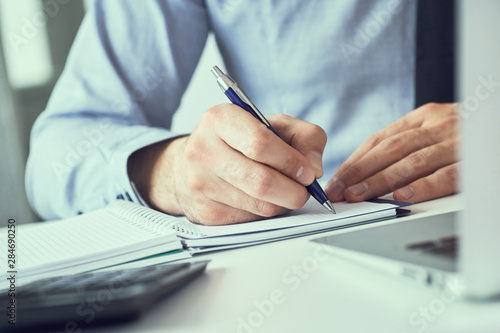 Businessman hand writing note on a notebook. Business man working at office desk. Close up of empty notebook on a blackboard with office supplies.