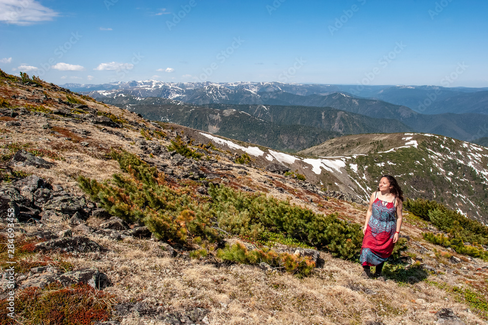 A European girl in a colored sundress stands on a mountainside with juniper bushes and smiles. The snow ridge goes into the distance. Sunny. Many mountain levels in the distance. 