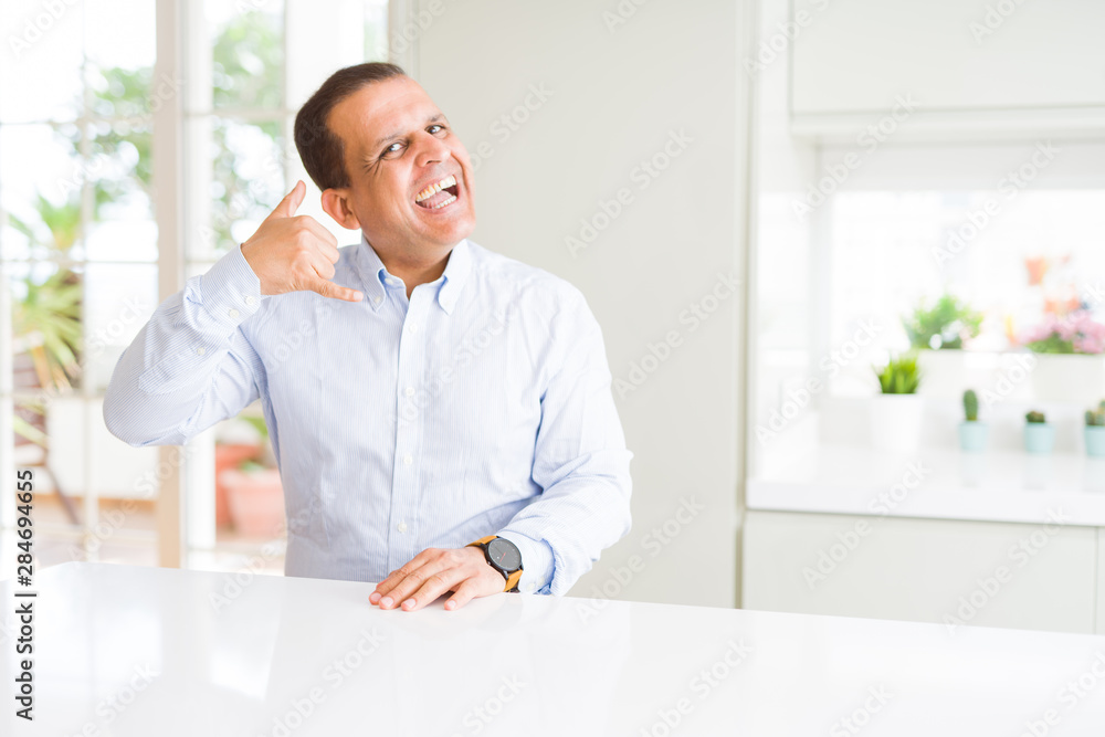 Middle age man sitting at home smiling doing phone gesture with hand and fingers like talking on the telephone. Communicating concepts. Middle age man sitting at home smiling doing phone gesture with hand and fingers like talking on the telephone. Communicating concepts.