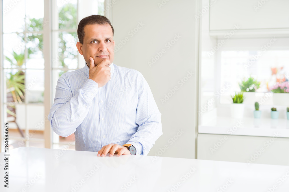 Middle age man sitting at home looking confident at the camera with smile with crossed arms and hand raised on chin. Thinking positive.