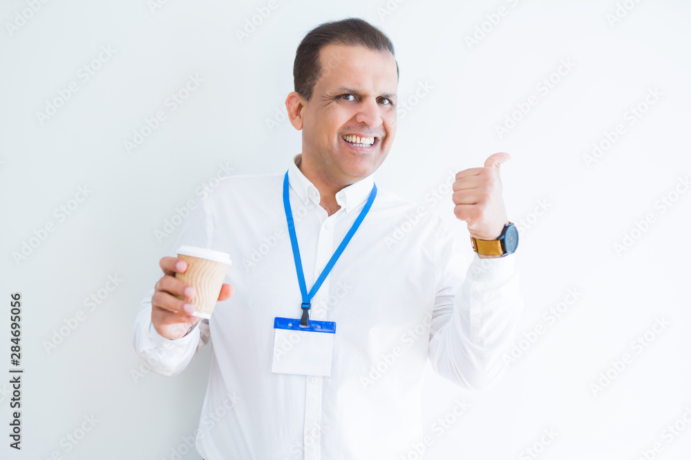 Middle age business man wearing ID card and drinking coffeeover white background pointing and showing with thumb up to the side with happy face smiling Middle age business man wearing ID card and drinking coffeeover white background pointing and showing with thumb up to the side with happy face smiling