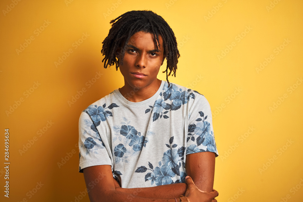 Afro man with dreadlocks wearing summer floral t-shirt over isolated yellow background skeptic and nervous, disapproving expression on face with crossed arms. Negative person. Afro man with dreadlocks wearing summer floral t-shirt over isolated yellow background skeptic and nervous, disapproving expression on face with crossed arms. Negative person.