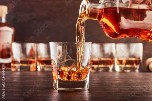 A glass of whiskey on a wooden table in which poured whiskey from a bottle. In the background are four glasses of whiskey, a full bottle of whiskey and a wooden bottle stopper.