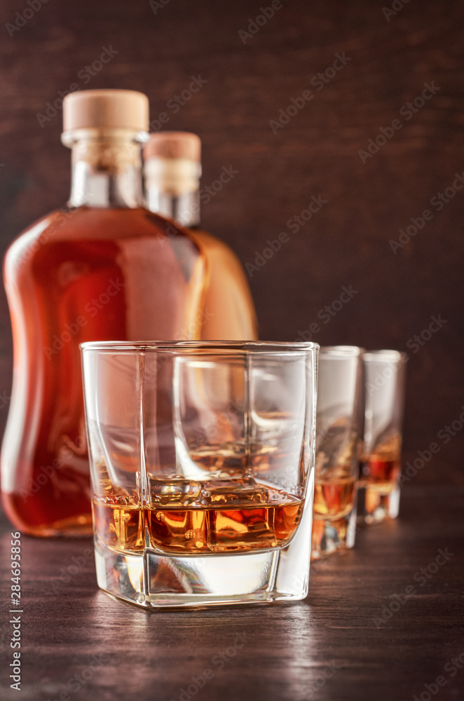 Three glasses of whiskey on the ice on the wooden table. In the background, two bottles of whiskey of different shapes. Three glasses of whiskey on the ice on the wooden table. In the background, two bottles of whiskey of different shapes.