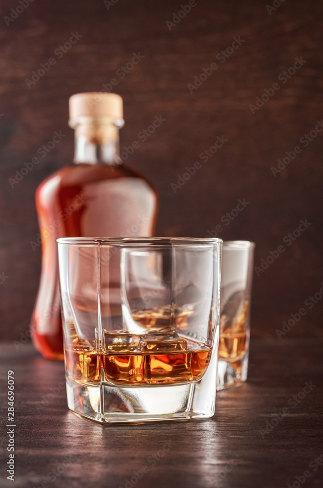 Two glasses of whisky with ice on a wooden table. There's a full bottle of whiskey in the background. Two glasses of whisky with ice on a wooden table. There's a full bottle of whiskey in the background.