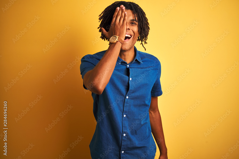 Afro man with dreadlocks wearing casual denim shirt standing over isolated yellow background covering one eye with hand, confident smile on face and surprise emotion. Afro man with dreadlocks wearing casual denim shirt standing over isolated yellow background covering one eye with hand, confident smile on face and surprise emotion.