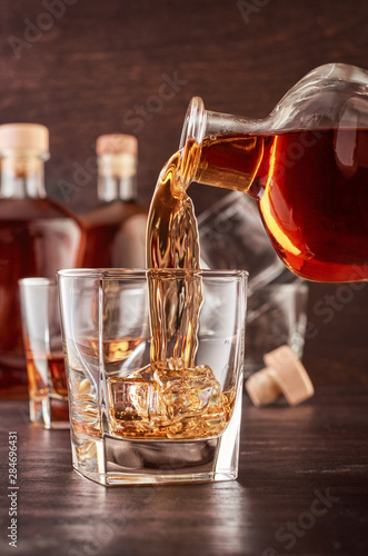 A glass of whiskey on a wooden table. In a glass filled with alcohol from the bottle. In the background, two bottles of whiskey of different shapes, a glass of whiskey and empty glasses of whiskey.