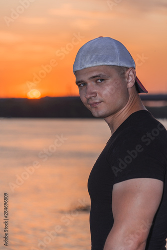 A young handsome tanned sports man in a cap stands on a sunset background with place for text. © Ирина Швейн