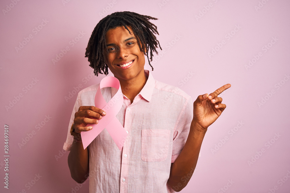 Afro american man with dreadlocks holding cancer ribbon over isolated pink background very happy pointing with hand and finger to the side Afro american man with dreadlocks holding cancer ribbon over isolated pink background very happy pointing with hand and finger to the side