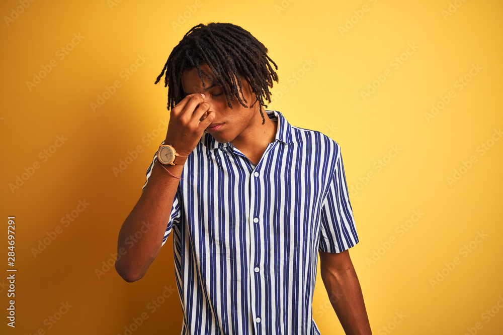 Afro man with dreadlocks wearing casual striped t-shirt over isolated yellow background tired rubbing nose and eyes feeling fatigue and headache. Stress and frustration concept. Afro man with dreadlocks wearing casual striped t-shirt over isolated yellow background tired rubbing nose and eyes feeling fatigue and headache. Stress and frustration concept.