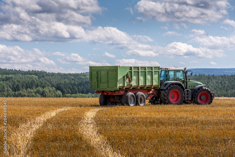 Tractor and trailer driving over a farm field leaving a tyre track in the dried grass on a cloudy blue sky day Tractor and trailer driving over a farm field leaving a tyre track in the dried grass on a cloudy blue sky day
