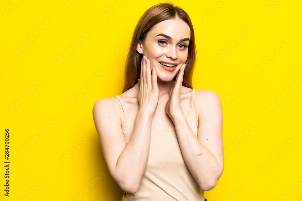 Shocked brunette woman holding her cheeks and looking at the camera with open mouth over yellow background Shocked brunette woman holding her cheeks and looking at the camera with open mouth over yellow background