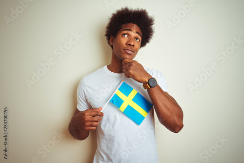 Young afro american man holding Sweden Swedish flag standing over isolated white background serious face thinking about question, very confused idea