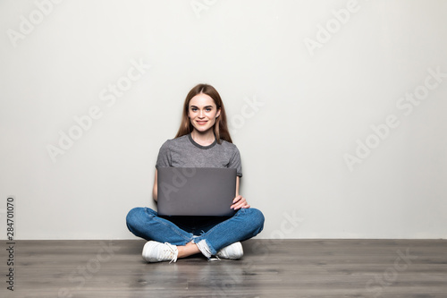 Attractive young woman holding laptop computer while sitting on the floor with legs crossed and looking away at copy space isolated over gray background © dianagrytsku