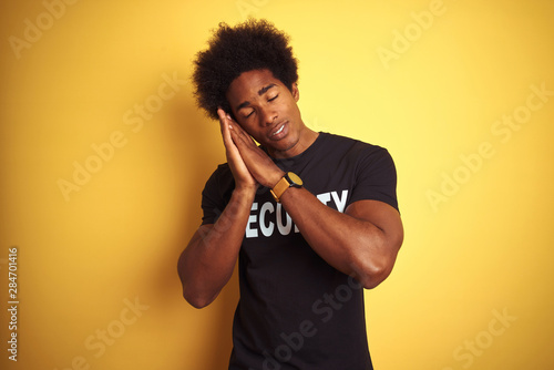 American safeguard man with afro hair wearing security uniform over isolated yellow background sleeping tired dreaming and posing with hands together while smiling with closed eyes. © Krakenimages.com