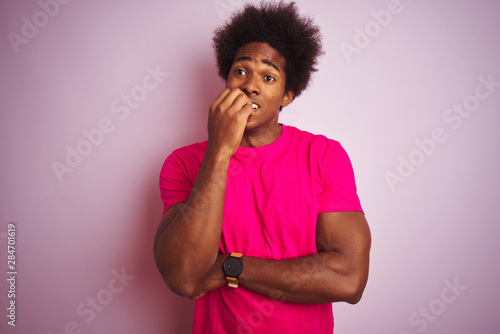 Young american man with afro hair wearing t-shirt standing over isolated pink background looking stressed and nervous with hands on mouth biting nails. Anxiety problem. © Krakenimages.com