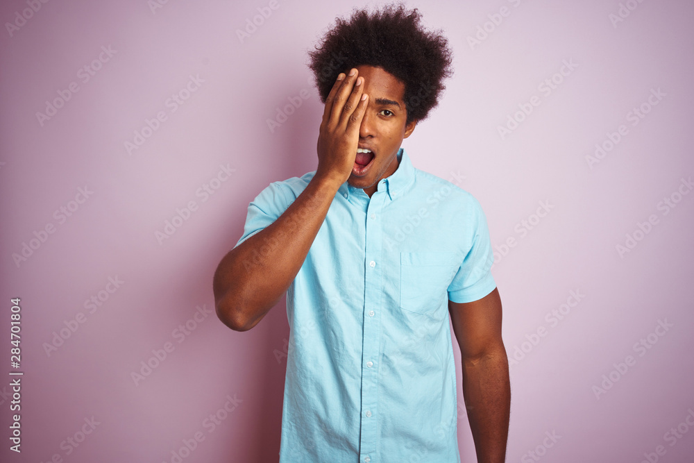 Young american man with afro hair wearing blue shirt standing over isolated pink background covering one eye with hand, confident smile on face and surprise emotion. Young american man with afro hair wearing blue shirt standing over isolated pink background covering one eye with hand, confident smile on face and surprise emotion.