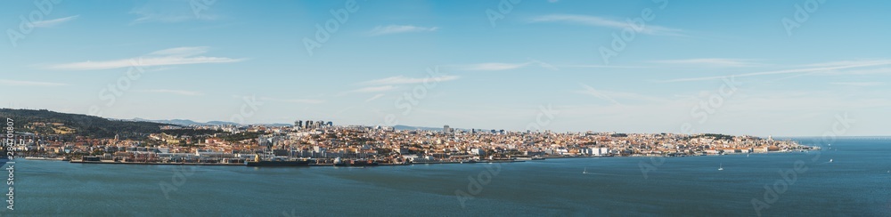 Panoramic view of Lisbon, Portugal from the Tagus river: waterline in the foreground, long river bank with a commercial docks near water, plenty of old houses and districts in the background