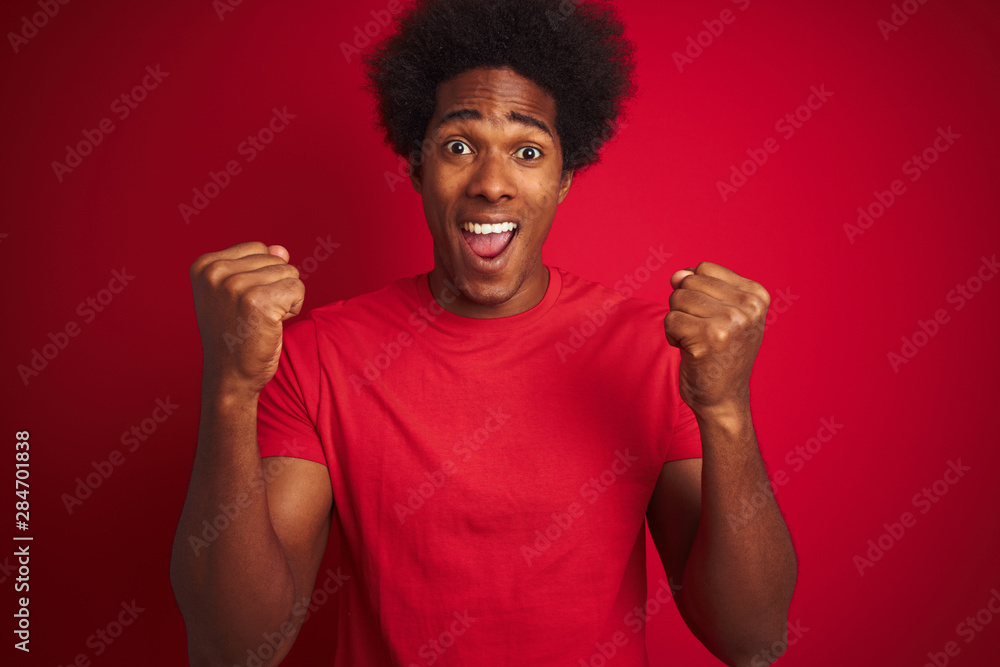 Young american man with afro hair wearing t-shirt standing over isolated red background celebrating surprised and amazed for success with arms raised and open eyes. Winner concept. Young american man with afro hair wearing t-shirt standing over isolated red background celebrating surprised and amazed for success with arms raised and open eyes. Winner concept.