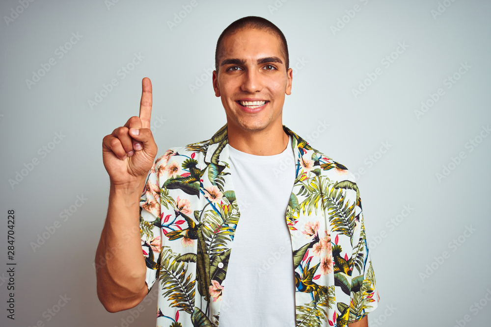 Young handsome man on holidays wearing Hawaiian shirt over white background showing and pointing up with finger number one while smiling confident and happy. Young handsome man on holidays wearing Hawaiian shirt over white background showing and pointing up with finger number one while smiling confident and happy.