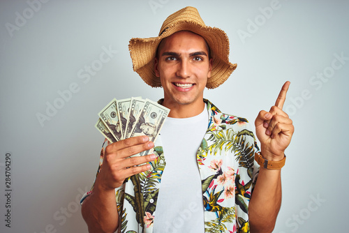 Young man on holidays holding dollars bank notes over white isolated background very happy pointing with hand and finger to the side