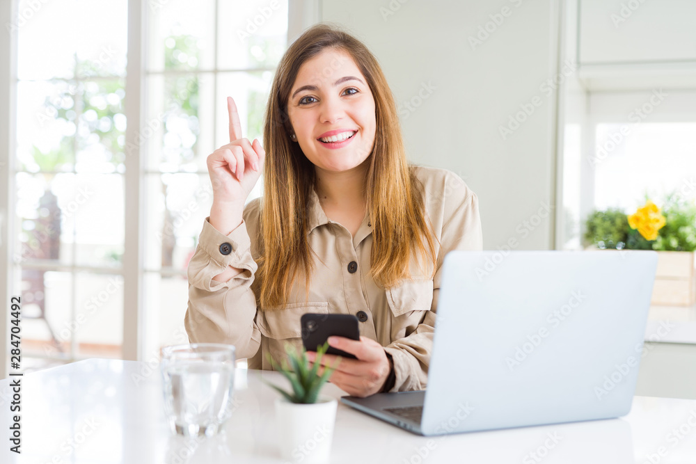 Beautiful young woman using smartphone and computer surprised with an idea or question pointing finger with happy face, number one Beautiful young woman using smartphone and computer surprised with an idea or question pointing finger with happy face, number one