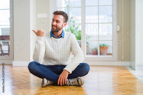 Handsome man wearing casual sweater sitting on the floor at home smiling with happy face looking and pointing to the side with thumb up.