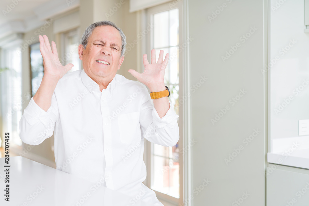 Fototapeta premium Handsome senior man at home crazy and mad shouting and yelling with aggressive expression and arms raised. Frustration concept.