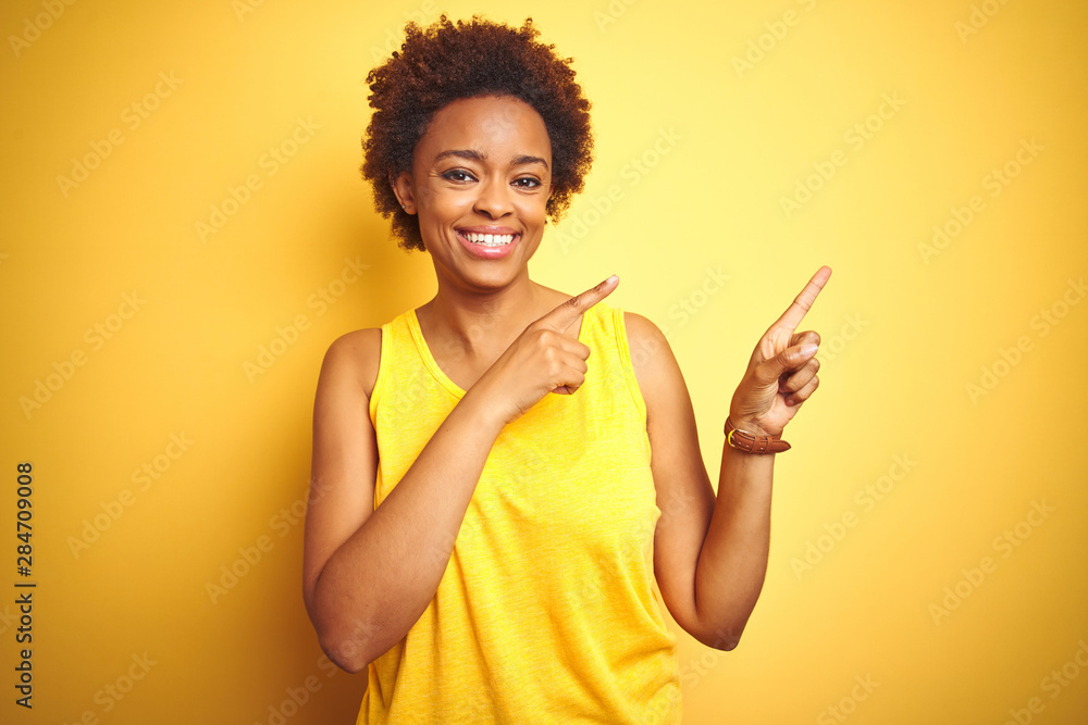Beauitul african american woman wearing summer t-shirt over isolated yellow background smiling and looking at the camera pointing with two hands and fingers to the side. Beauitul african american woman wearing summer t-shirt over isolated yellow background smiling and looking at the camera pointing with two hands and fingers to the side.