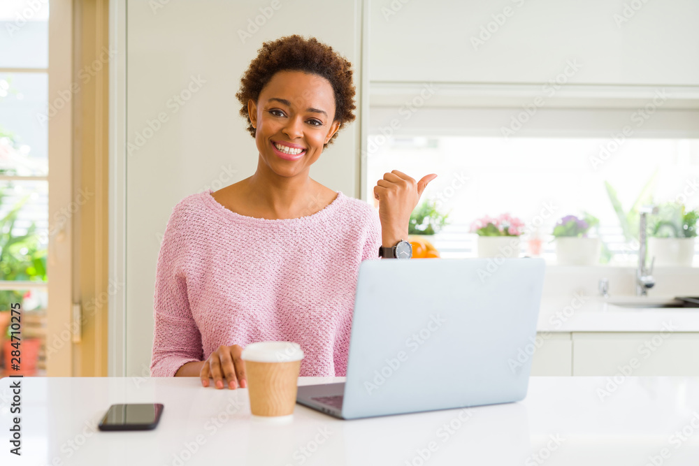 Young african american woman working using computer laptop smiling with happy face looking and pointing to the side with thumb up.