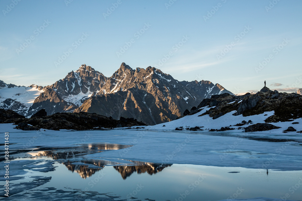 © DCrane Photography - Spring thaw on remote alpine lake in the Talkeetna Mountains of Alaska. Man standing on ridge reflected in the lake.