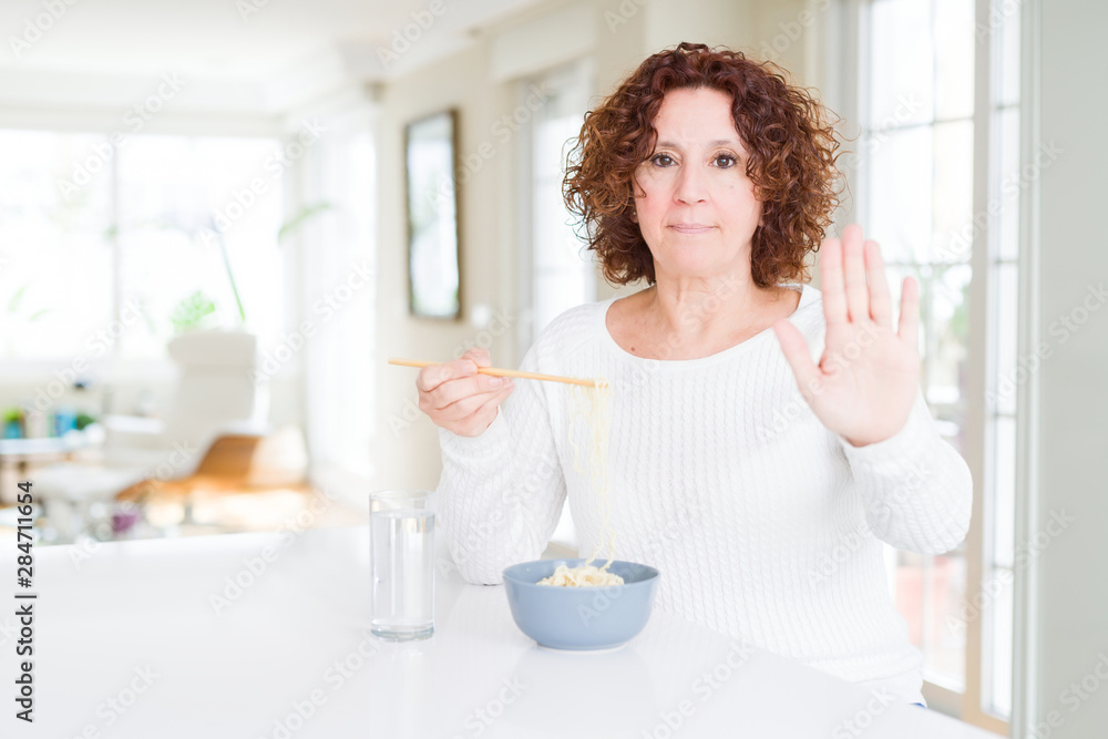 Senior woman eating asian noodles using chopsticks with open hand doing stop sign with serious and confident expression, defense gesture Senior woman eating asian noodles using chopsticks with open hand doing stop sign with serious and confident expression, defense gesture