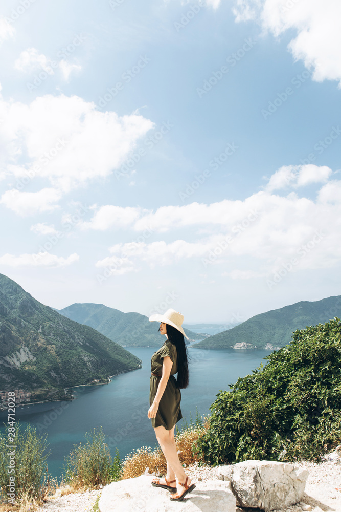Naklejka premium A girl in a hat on the background of the sea and mountains admires a beautiful view of the natural landscape in Montenegro.