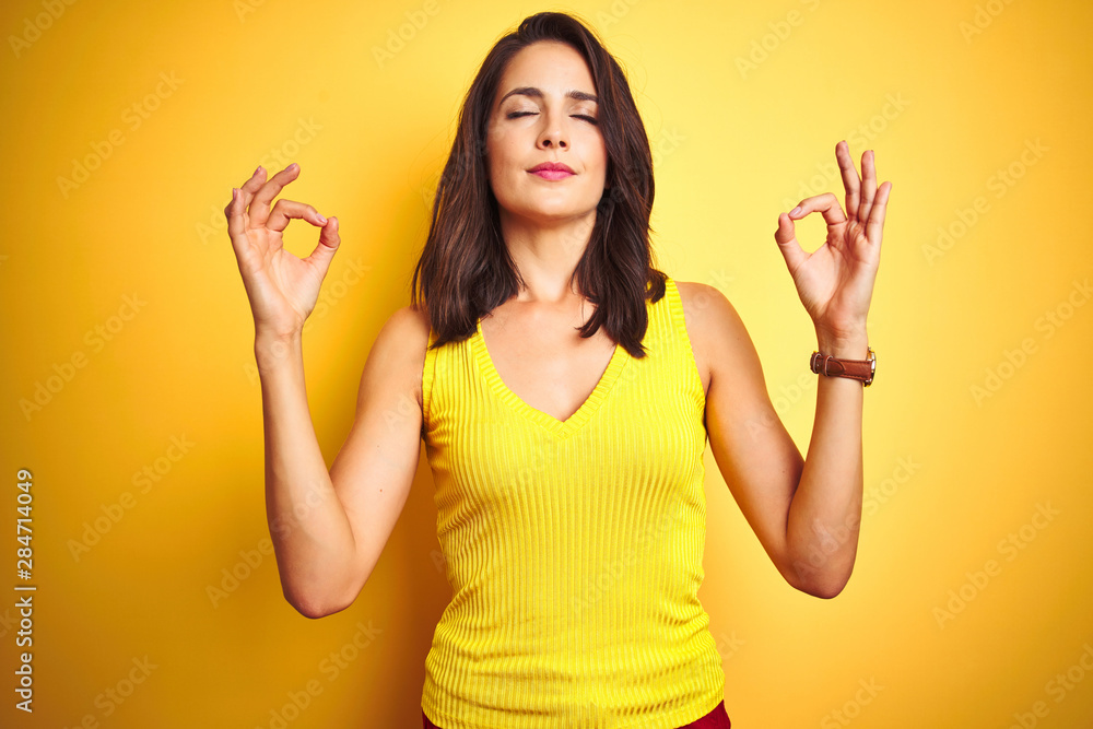 Young beautiful woman wearing t-shirt standing over yellow isolated background relax and smiling with eyes closed doing meditation gesture with fingers. Yoga concept. Young beautiful woman wearing t-shirt standing over yellow isolated background relax and smiling with eyes closed doing meditation gesture with fingers. Yoga concept.