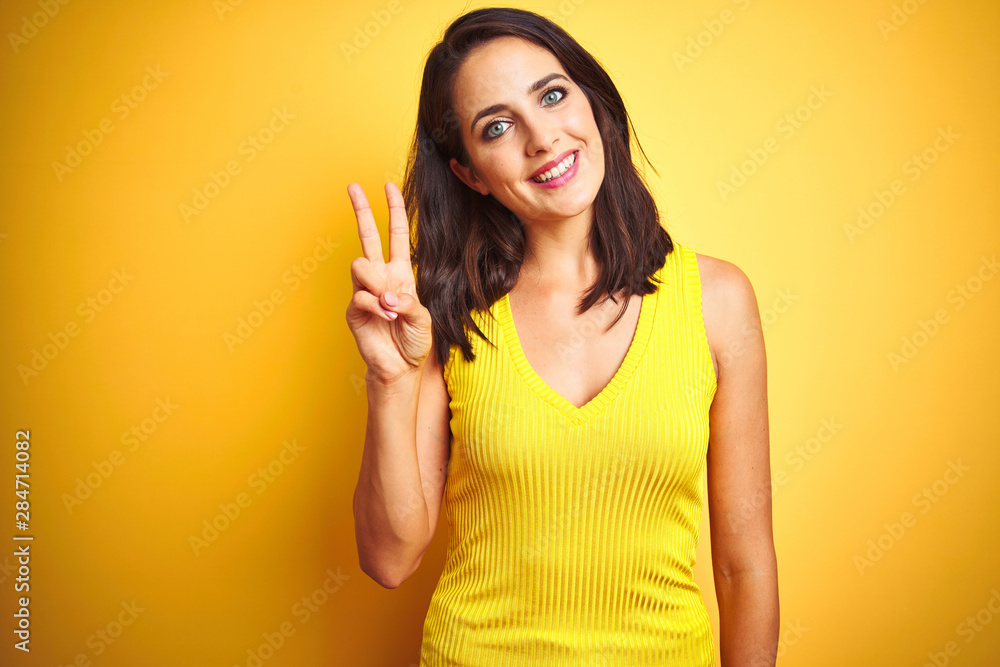 Young beautiful woman wearing t-shirt standing over yellow isolated background showing and pointing up with fingers number two while smiling confident and happy.