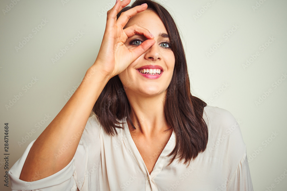 Young beautiful woman wearing casual shirt standing over white isolated background doing ok gesture with hand smiling, eye looking through fingers with happy face. Young beautiful woman wearing casual shirt standing over white isolated background doing ok gesture with hand smiling, eye looking through fingers with happy face.