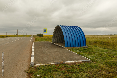 empty blue bus stop on the road against a cloudy sky © Mieszko9