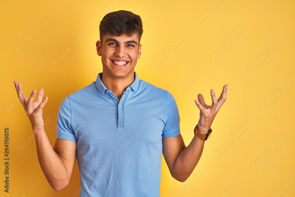 Young indian man wearing blue polo standing over isolated yellow background celebrating mad and crazy for success with arms raised and closed eyes screaming excited. Winner concept Young indian man wearing blue polo standing over isolated yellow background celebrating mad and crazy for success with arms raised and closed eyes screaming excited. Winner concept