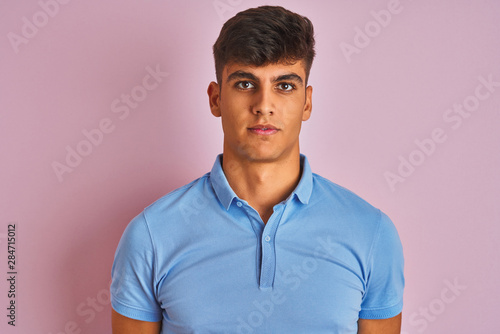 Young indian man wearing blue polo standing over isolated pink background with serious expression on face. Simple and natural looking at the camera. © Krakenimages.com