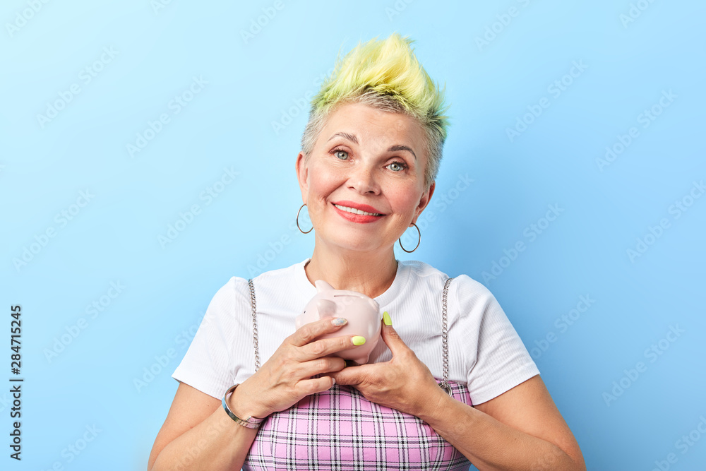 funny cool old granny with dyed yellow hair and fashion clothes holding  piggy bank and posing to the camera. close up portrait, isolated blue  background, studio shot. woman putting her pension foto