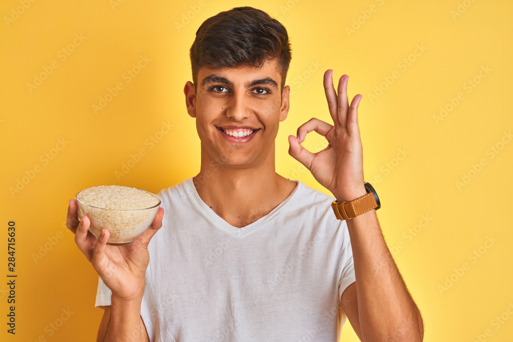 Young indian man holding bowl with rice standing over isolated yellow background doing ok sign with fingers, excellent symbol Young indian man holding bowl with rice standing over isolated yellow background doing ok sign with fingers, excellent symbol